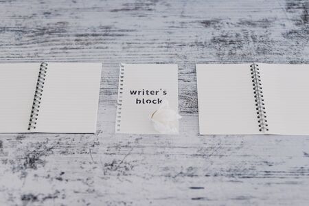 creative block or lack of inspiration conceptual still-life, piece of paper on wooden desk with Writer's Block text surrounded by blank notepads and with scrunched paper ball next to itの写真素材