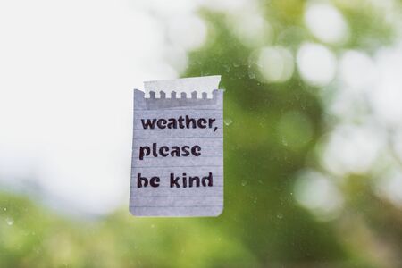 weather events and climate change conceptual still-life, Weather please be kind message with sticky tape on window glass with bokeh on stormy weather and grey sky over a backyardの写真素材