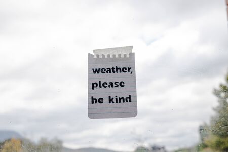 weather events and climate change conceptual still-life, Weather please be kind message with sticky tape on window glass with bokeh on stormy weather and grey sky over a backyardの写真素材