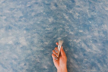 hand holding brush on top of unfinished work of an abstract textured painting or photography backdrop with blue and white toned acrylic paintの写真素材