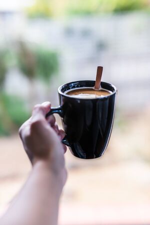 hand holding mug of warm coffee in front of window with backyard background shot at shallow depth of fieldの写真素材