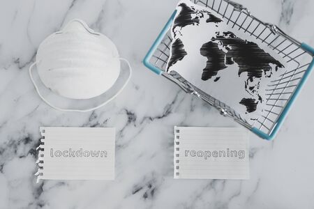 going back to normal after the global pandemic of covid-19, lockdown versus reopening signs next to shopping cart with world map and mask with glovesの写真素材