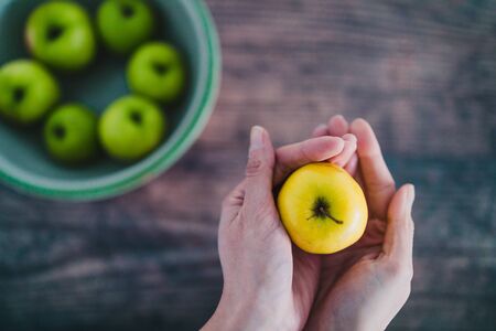 healthy plant-based food ingredients concept, bowl of green apples and hand holding a smaller yellow one next to itの写真素材