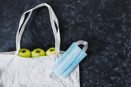 coronavirus disposable mask on top of reusable grocery bag with green apples in it, concept of...