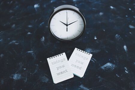 meditation and psychology concept, notepads with titles Things you need vs things you want side by side on desk next to clock metaphor of the present momentの写真素材