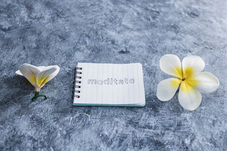 self-care and mental health concept, notepad with text Meditate next to tropical flowers on grey concrete surfaceの写真素材