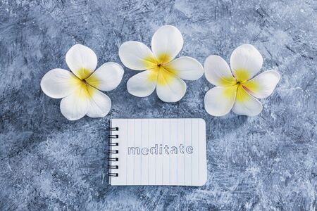 self-care and mental health concept, notepad with text Meditate next to tropical flowers on grey concrete surfaceの写真素材