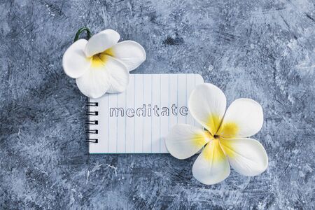 self-care and mental health concept, notepad with text Meditate next to tropical flowers on grey concrete surfaceの写真素材