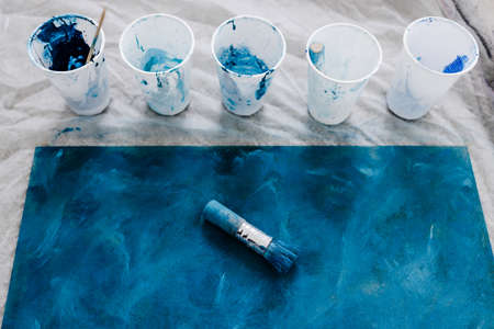 arts and craft hobbies concept, blue and black acrylic painting with colors in plastic cups and mixed painting tools next to it while the work is in progressの写真素材