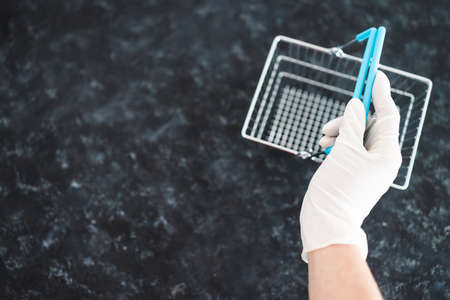 the new normal after the covid-19 virus pandemic outbreak, hand with disposable gloves holding miniature supermarket shopping cart not to touch potentially infected surfacesの写真素材