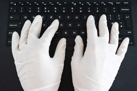 the new normal after thecovid-19 virus pandemic outbreak, hands typing on shared computer keyboard at work wearing disposable gloves to avoid contact with potentially infected surfacesの写真素材