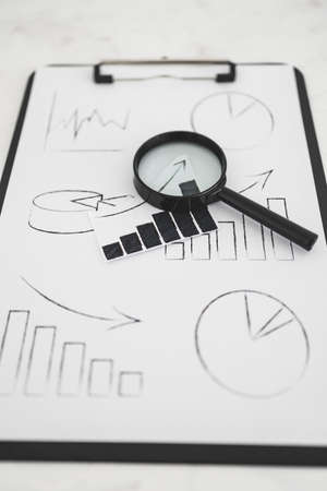 business analysis and statistics concept, desk with mixed graphs and charts on clipboard with magnifying glass on itの写真素材