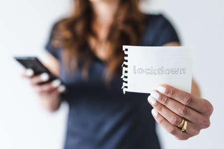 life after the covid-19 pandemic outbreak, woman holding Lockdown text towards the camera while browsing on her smartphone shot at shallow depth of fieldの写真素材