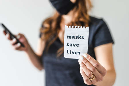 life after the covid-19 pandemic outbreak, woman holding Masks save lives text towards the camera while browsing on her smartphone shot at shallow depth of fieldの写真素材