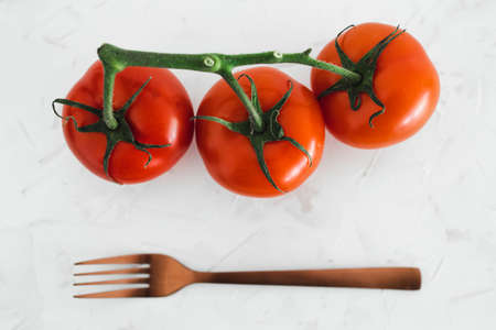 simple ingredients and whole food diet concept, close-up of three tomatoes on vine with fork next to them on white minimalist backgroundの写真素材