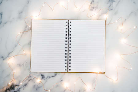 blank notebook with pages open on marble desk surrounded by fairy lights with copy space to add your...