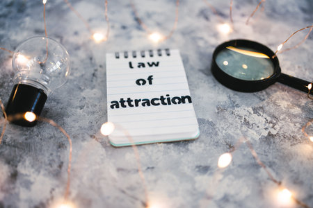 law of attraction notepad with text next to light bulb and magnifying glass symbol of introspection and mindset shift surrounded by fairy lightsの写真素材