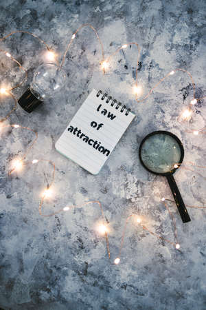 law of attraction notepad with text next to light bulb and magnifying glass symbol of introspection and mindset shift surrounded by fairy lightsの写真素材
