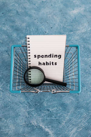 spending habits text on notepad with shopping basket and magnifying glass, money and consumer...