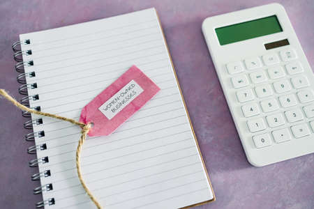 women-owned business tag with calculator and notepad on pink desk, supporting equality and equal...