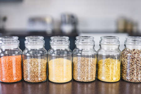 clear pantry jars with different types of grains and legumes including quinoa  lentils buckwheat and barley on benchtop with kitchen bokeh, simple ingredients conceptの写真素材