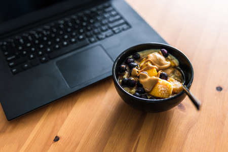eating mango blueberries oatmeal while working from home on a laptop, concept of remote working or lunch at the officeの写真素材