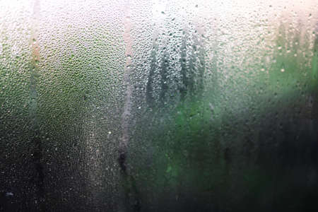 water droplets of humidity condensation on window glass seen from indoor with backyard bokeh in the...