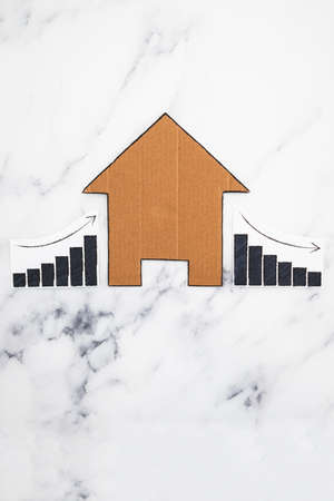 mortgages rentals and property prices increasing and decreasing, house icon made of cardboard with graph showing stats going up and down conceptual image on marble backgroundの写真素材