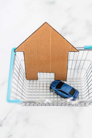 getting a car and home loan or choosing investments and insurance conceptual image, house icon and toy car inside of shopping basket on white marble backgroundの写真素材