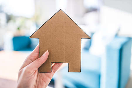 hand holding house icon in front of living room bokeh, concept of buying a house and real estate market or dream home or family lifeの写真素材