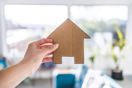 hand holding house icon in front of living room bokeh, concept of buying a house and real estate market or dream home or family lifeの写真素材