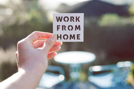 work from home sign being hold in front of sunny out of focus backard with table and chairs, concept of digital nomads working remotely or wfh days during lockdowns or covid-19 isolationの写真素材