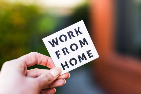 work from home sign being hold in front of out of focus lush green backard and home exterior,...