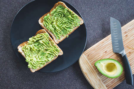 basic plant-based smashed avocado toast in the making on kitchen counter with half avocado and...