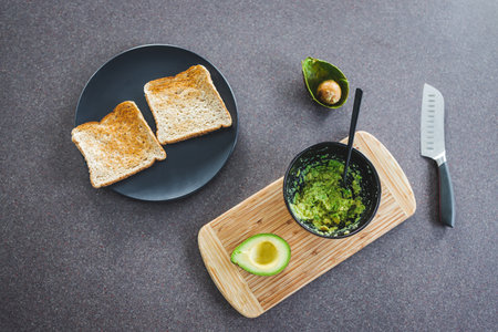 basic plant-based smashed avocado toast in the making on kitchen counter with half avocado and...