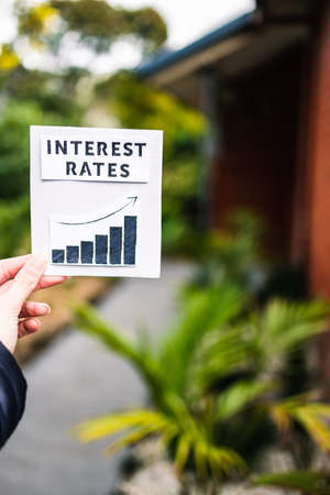 property affordabilikty and home loan payment increases, hand holding interest rates sign with graph showing stats going up in front of backyard and home exterior bokehの写真素材