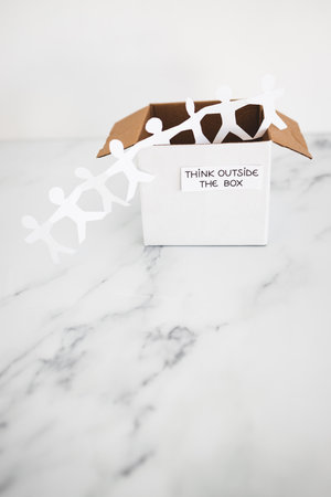 Think outside the box concept with paper people chain getting out of a white box with text on white marble	with minimalistic compositionの写真素材