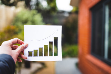 property value and return on investment or rent price conceptual image, hand holding real estate graph showing stats going up with cut outs in front of house exterior and backyardの写真素材