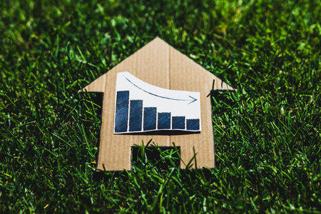 interest rates going down, graph showing stats decreasing over house icon made of cardboard on green lawnの写真素材