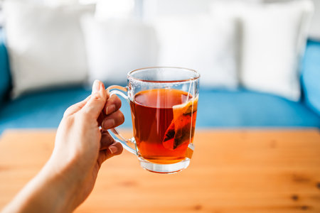 hand holding a cup of rooibos tea with teabag on coffee table in front of blue couch with white cushionsの写真素材