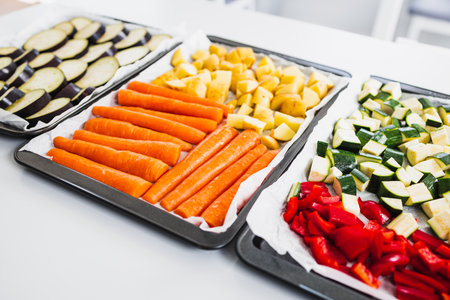 chopped carrots, potatoes, zucchini, capsicum, and eggplant on baking trays ready for roasting, a colorful healthy meal prep sceneの写真素材