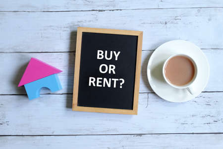 Top view of house model and blackboard written with ' BUY OR RENT?' with cup of coffee on white wooden background. Real estate,business and finance theme.の写真素材