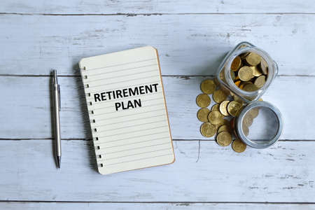 Top view of notebook written with 'RETIREMENT PLAN' with pen and coins on white wooden background.の写真素材