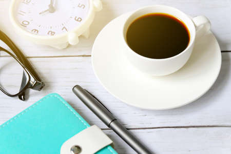 Top view and selective focus of a cup of black coffee with table clock,sunglasses, notebook and pen on white wooden background.の写真素材