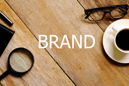 Top view of a cup of coffee,eyeglasses,magnifying glass,notebook and pen on wooden background written with BRAND.の写真素材