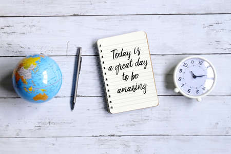 Top view of globe of world map,table clock,pen and notebook written with inspirational quotes 'TODAY IS A GREAT DAY TO BE AMAZING' on white wooden background.の写真素材