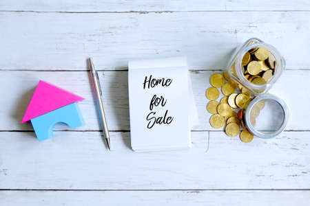 Top view of a jar of coins,home model,pen and notebook written with HOME FOR SALE on white wooden background.の写真素材