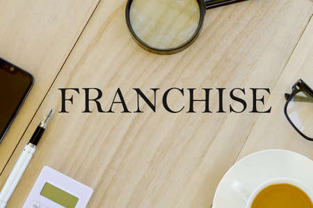 Business and finance concept Top view of magnifying glass,eyeglasses,a cup of coffee,calculator,pen and mobile phone on wooden background written with Franchise.の写真素材