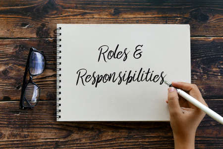 Top view of glasses and hand holding pen writing Roles and Responsibilities on notebook on wooden...