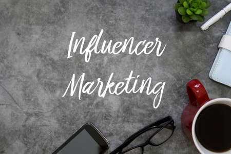 Top view of mobile phone,glasses,coffee,notebook,pen and plant on grey grunge floor written with Influencer Marketing.の写真素材